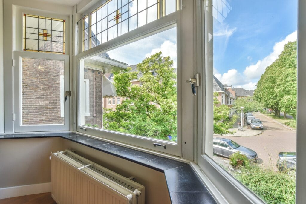 Interior view through open white casement windows with stained glass toplights, overlooking a tree-lined residential street