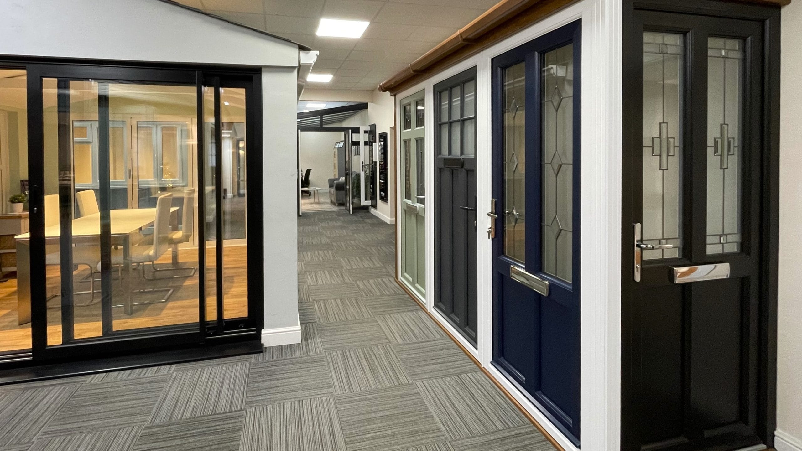 Interior of our showroom displaying composite doors and a black bi-fold door opening onto a furnished room display area.
