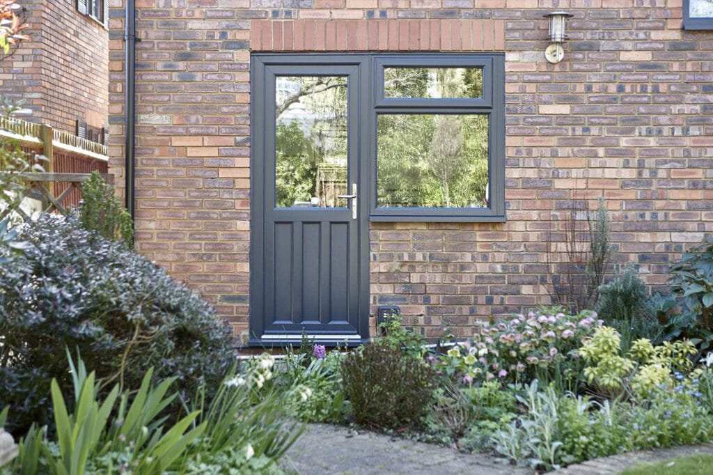 Anthracite grey uPVC door with large glazed panel and side window, set in red brick wall surrounded by cottage garden planting.