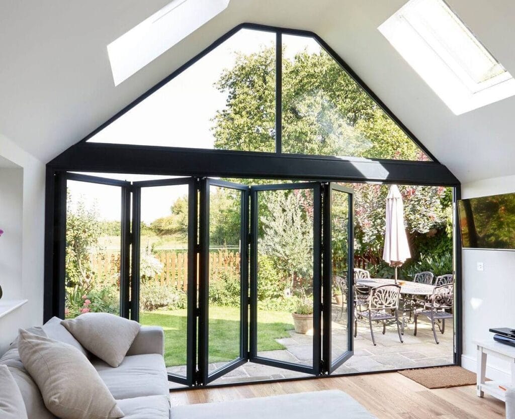 Anthracite grey aluminium bi-fold doors open onto a sunny garden patio, with a vaulted glazed gable and roof skylights above