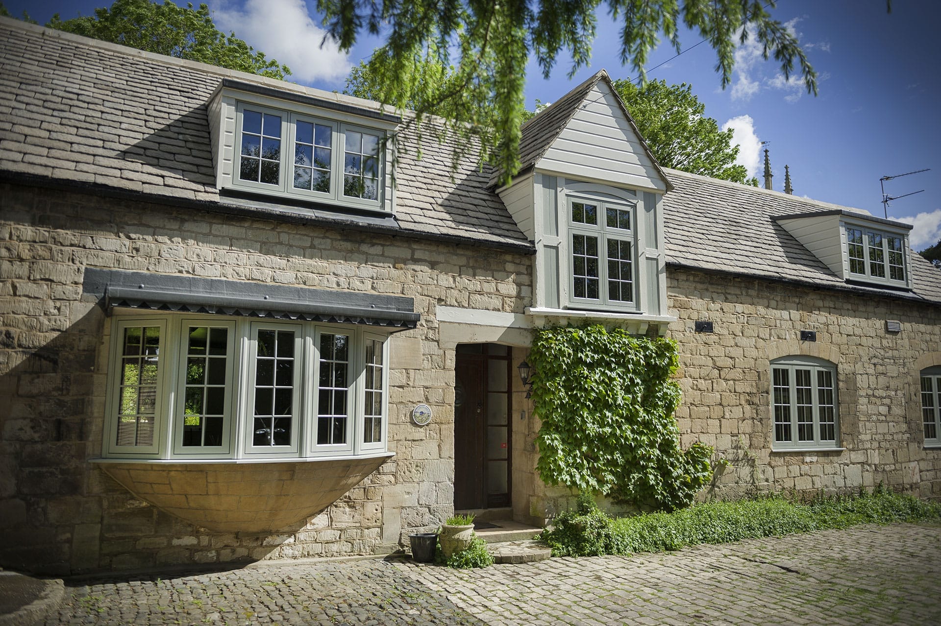 Stone cottage exterior with sage green casement windows, a bay window and dormers, set on a cobbled driveway