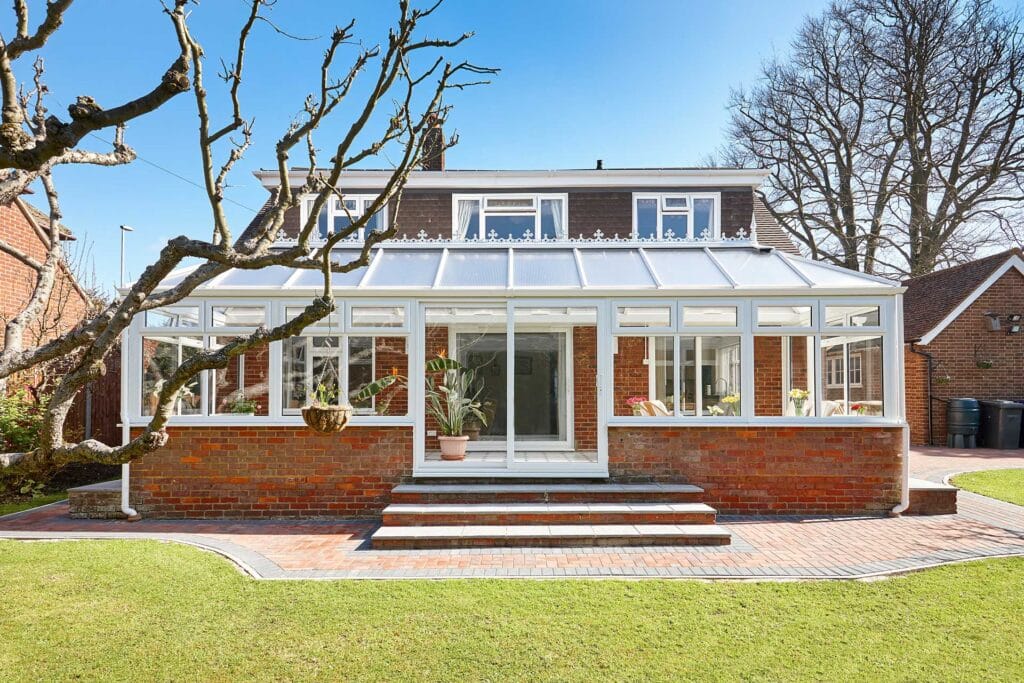 Large white uPVC lean-to conservatory with decorative ridge detail on the rear of a red brick detached house